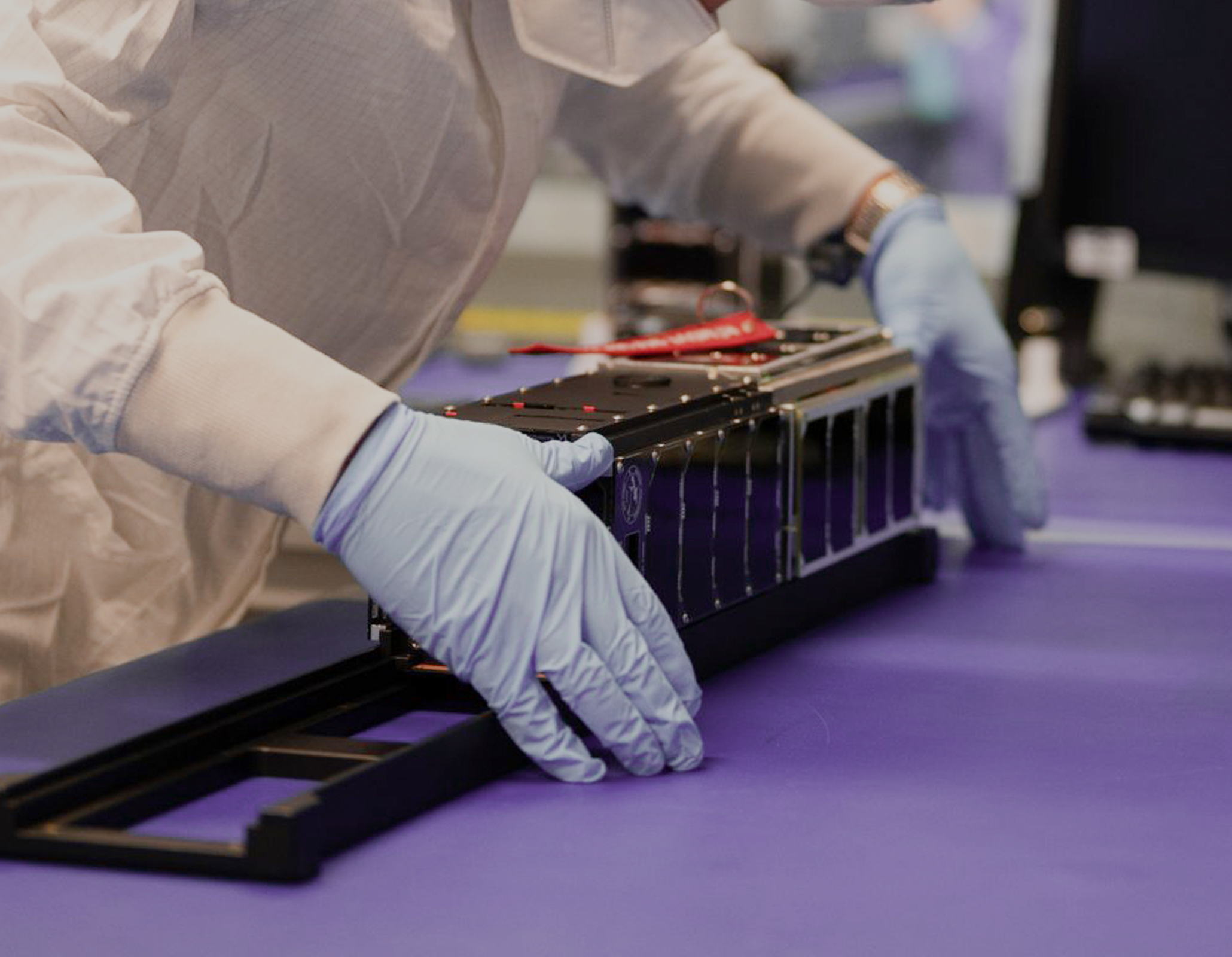 Technician handling satellite equipment in cleanroom environment.